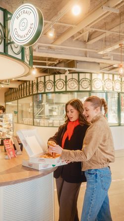 Two women choosing doughnuts at a counter inside Krispy Kreme - Gare de Montparnasse with a Retrans Commandes sign overhead.