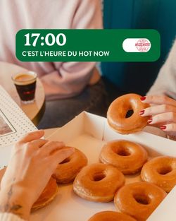 Hands reaching for glazed doughnuts in a Krispy Kreme box at a café table with coffee cups nearby.
