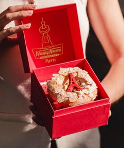 Doughnut décoré avec des amandes et une petite tour Eiffel, présenté dans une boîte rouge Krispy Kreme.