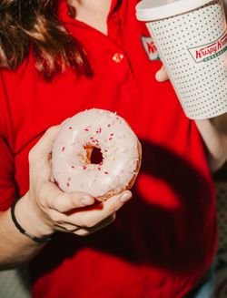 Hand holding a pink-frosted doughnut with sprinkles and a Krispy Kreme branded coffee cup in the other hand.