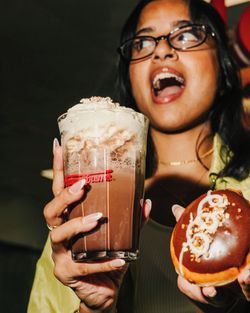 Woman holding a glass of whipped cream topped coffee and a chocolate glazed doughnut with white icing swirl