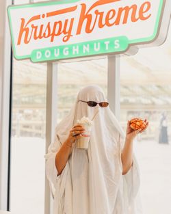 Personne déguisée avec un drap blanc tenant un café et un doughnut sous l'enseigne Krispy Kreme.