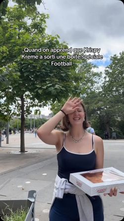 Jeune femme tenant une boîte de doughnuts Krispy Kreme en extérieur, avec des arbres et un ciel nuageux en arrière-plan.
