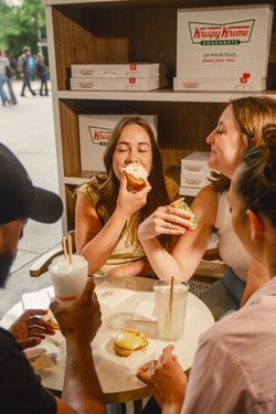 Quatre personnes dégustent des doughnuts et des boissons dans un cadre de café Krispy Kreme avec boîtes en arrière-plan.