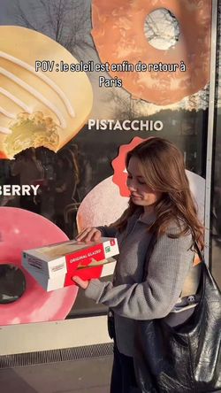 Une femme tenant une boîte de doughnuts devant la vitrine de Krispy Kreme Paris avec des images de pâtisseries.