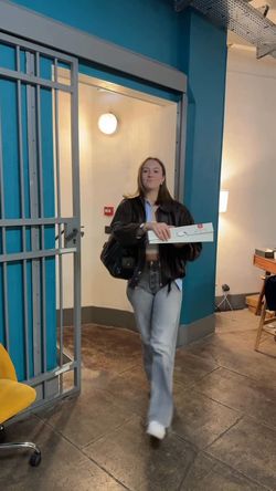 Une cliente portant une boîte de beignets sort d'un café, vêtue d'une veste en cuir et de jeans.