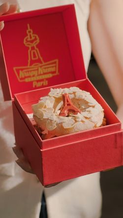 Doughnut décoré sur un fond rouge, présenté dans une boîte Krispy Kreme avec la Tour Eiffel.
