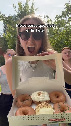 Boîte contenant six doughnuts variés, posée devant trois personnes avec des arbres en arrière-plan.