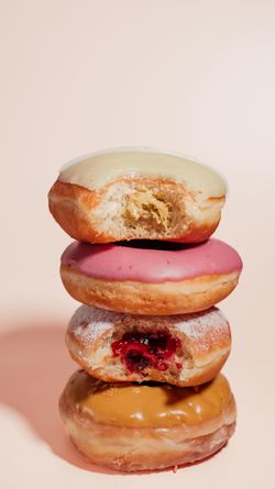 Quatre doughnuts empilés avec divers glaçages et garnitures visibles sur un fond rose clair.