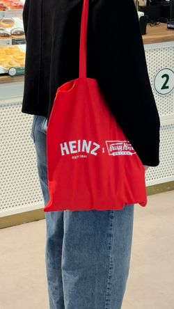 Red tote bag with Heinz and Krispy Kreme logos carried by a person in jeans and black top inside Krispy Kreme shop.