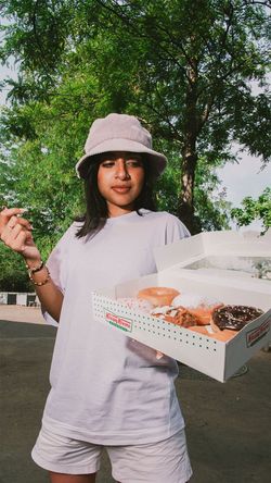 Boîte ouverte de doughnuts Krispy Kreme tenue par une femme en extérieur proche d'arbres sous lumière naturelle.