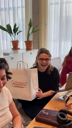 Krispy Kreme takeout bag held by a woman in a bright room with plants and people seated around a table.