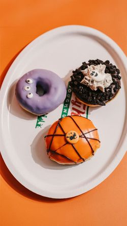 Trois doughnuts décorés avec du glaçage violet, orange et chocolat, présentés sur un plateau blanc chez Krispy Kreme.