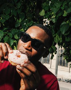 Homme portant des lunettes de soleil mangeant un doughnut glacé rose à l'extérieur près d'un arbre.