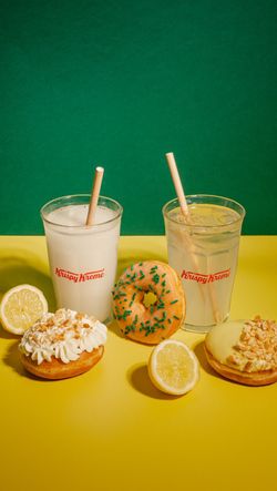 Three doughnuts with lemon toppings and two iced drinks with straws on a yellow surface and green background at Krispy Kreme.