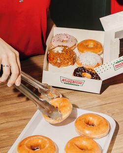 Boîte ouverte avec six doughnuts variés posée sur une table en bois, un salarié sert des doughnuts avec une pince.