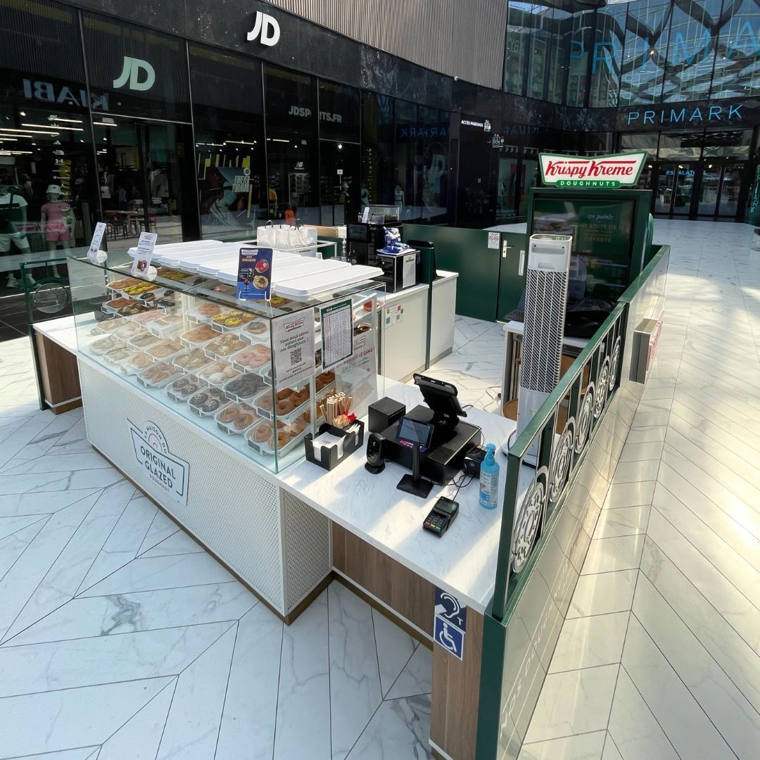 Clients montrant des boîtes de beignets devant un décor moderne au café, avec pâtisserie et ambiance conviviale.