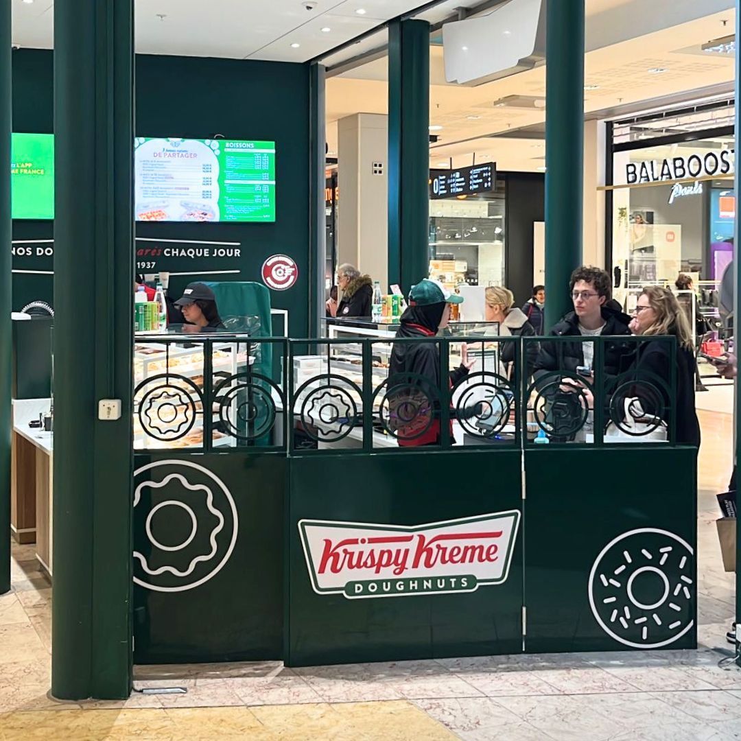 Stand Krispy Kreme avec des clients, offrant des doughnuts, café, et pâtisserie dans un cadre de centre commercial.